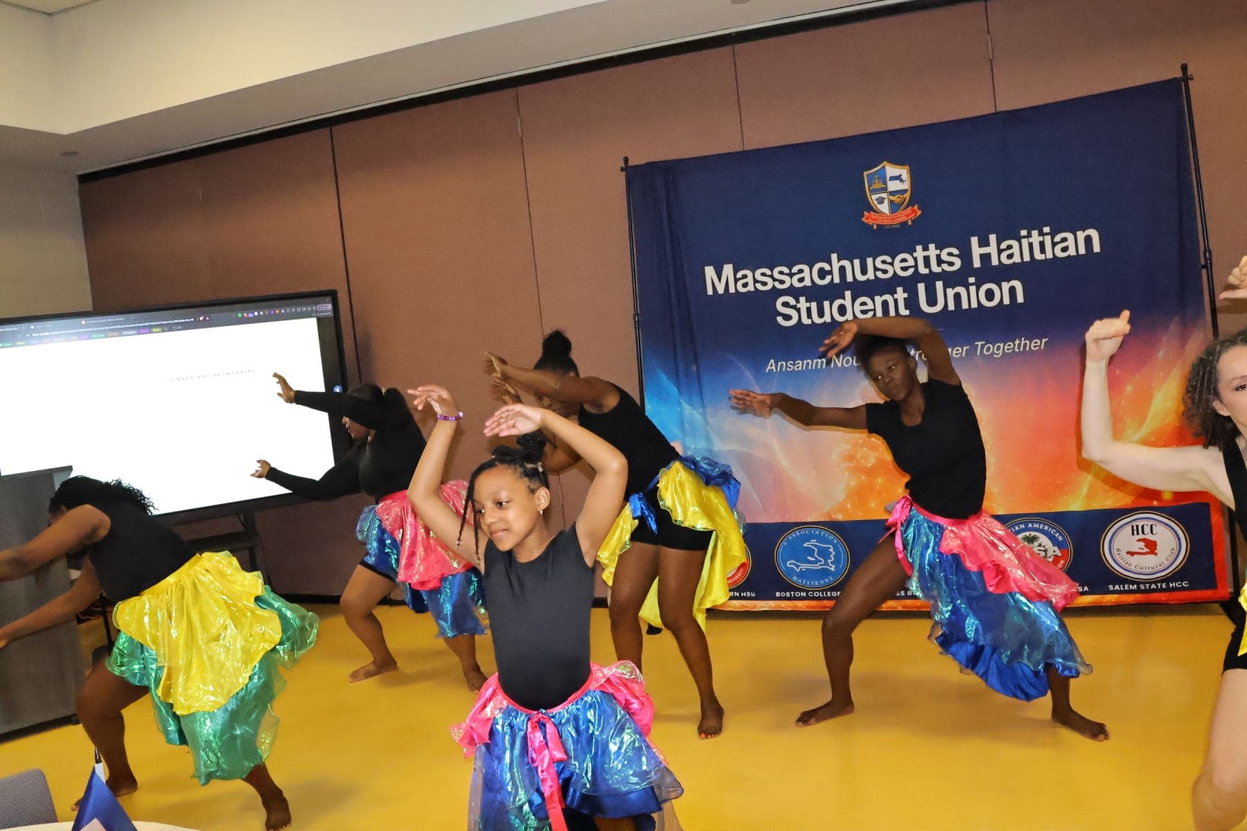 Young dancers in colorful skirts performing in front of the MAHSU banner
